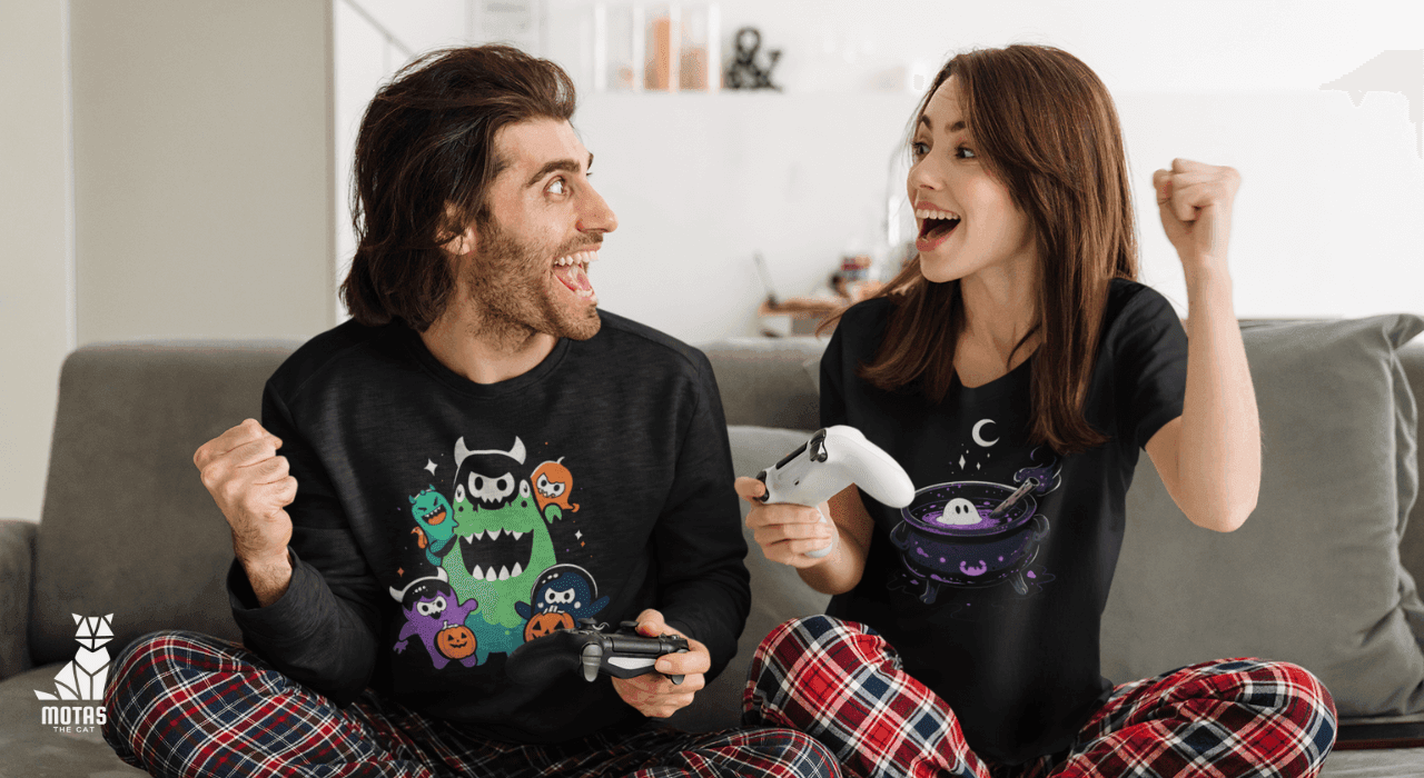 Excited couple in motasthecat gaming apparel playing video games on gray sofa, cheering with controllers in hand