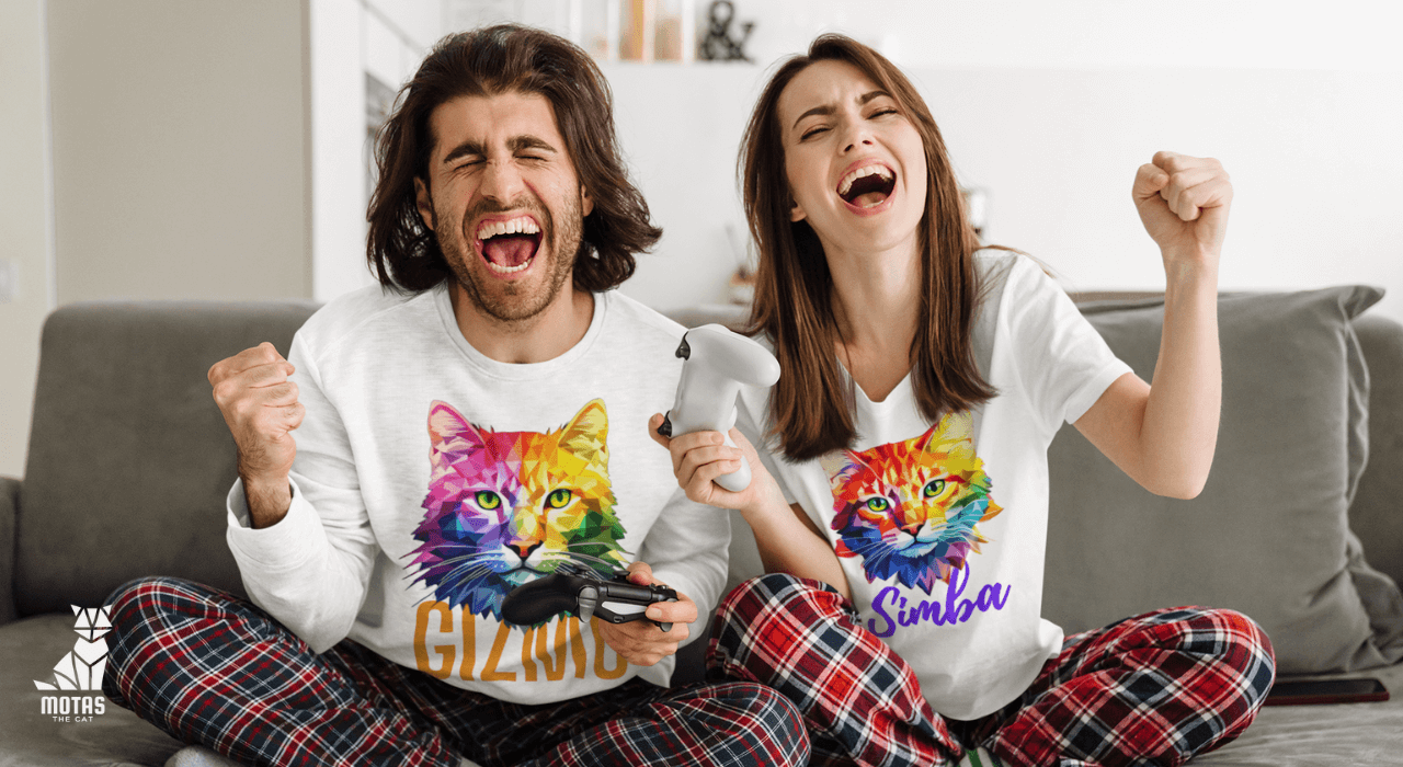 Excited couple wearing motasthecat The original pop culture t-shirts playing video games on gray sofa, celebrating victory with raised fists and game controllers